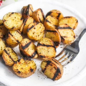 Grilled potato halves with visible char marks and herb seasoning on a white plate, with a fork placed on the side.