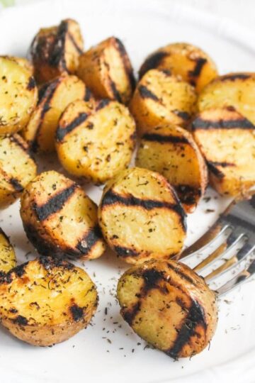 Grilled potato halves with visible char marks and herb seasoning on a white plate, with a fork placed on the side.