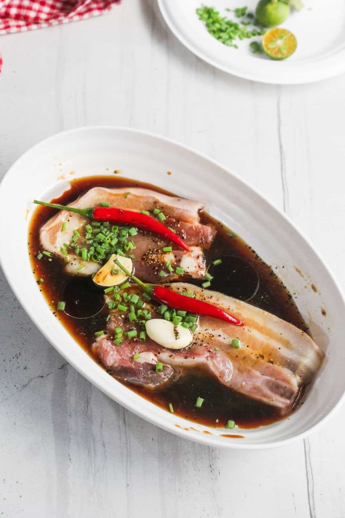 Two pieces of pork belly marinating in a dark sauce with whole red chilies, garlic cloves, and chopped green onions in a white oval dish on a white surface.