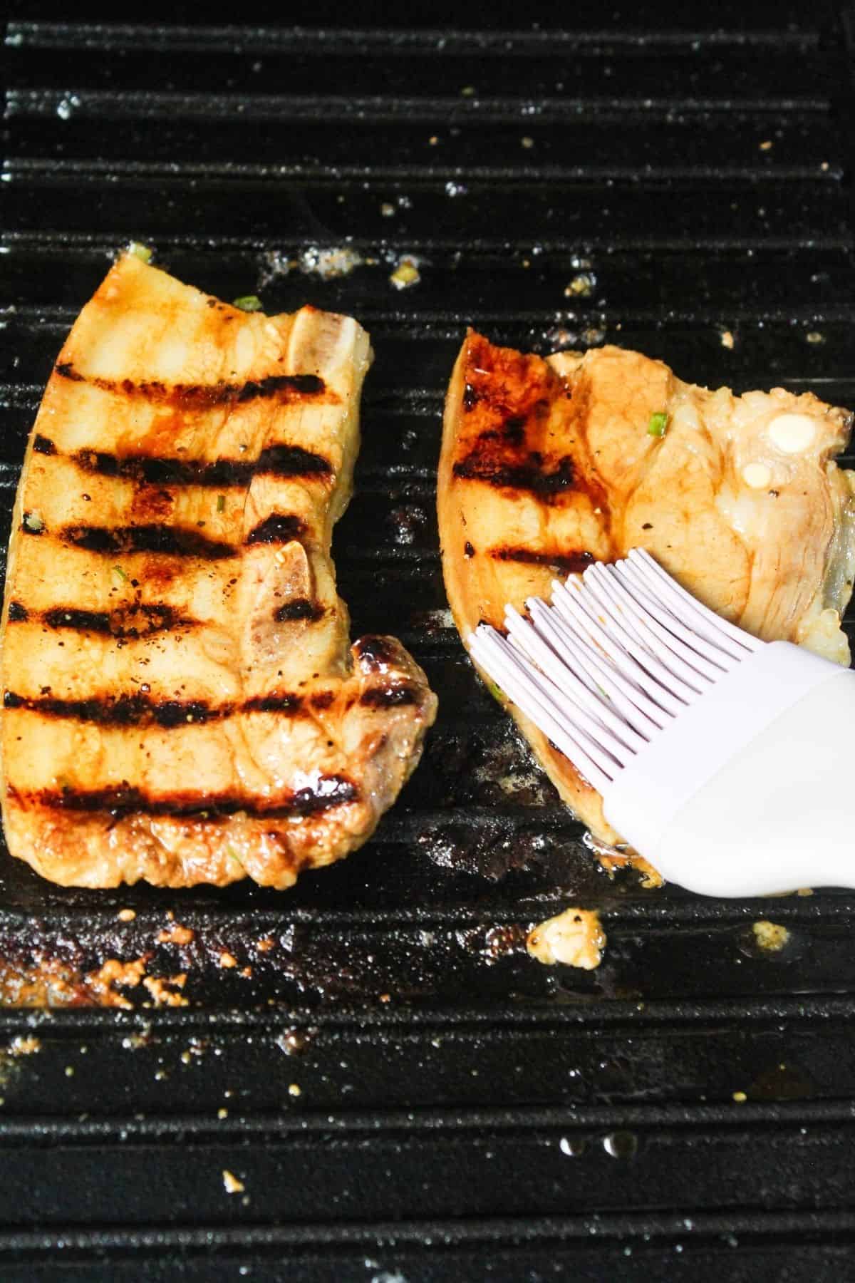 Two pieces of grilled meat on a grill pan, with a silicone brush applying marinade or sauce to one piece.