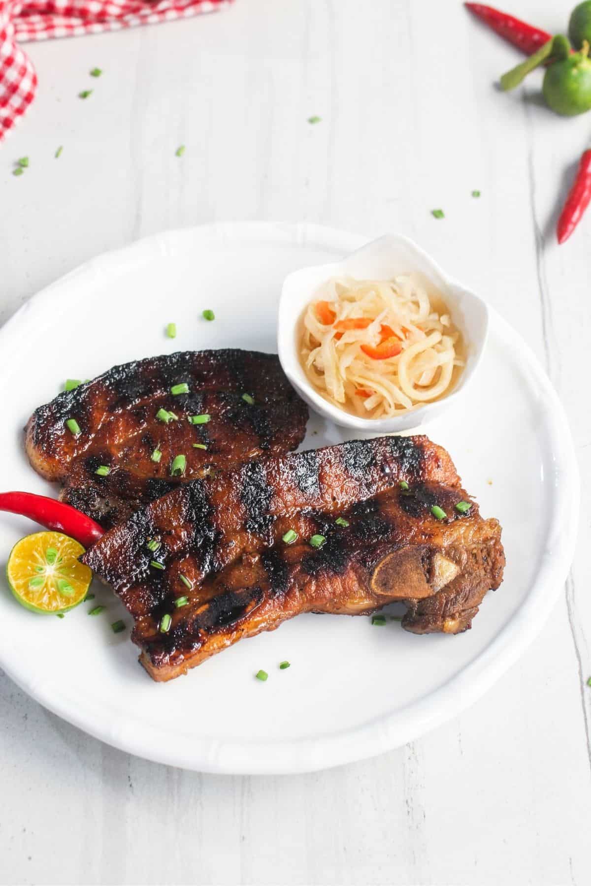 Grilled marinated pork chop on a white plate, garnished with chopped green onions, served with a small bowl of pickled vegetables, a red chili, and a sliced calamansi.