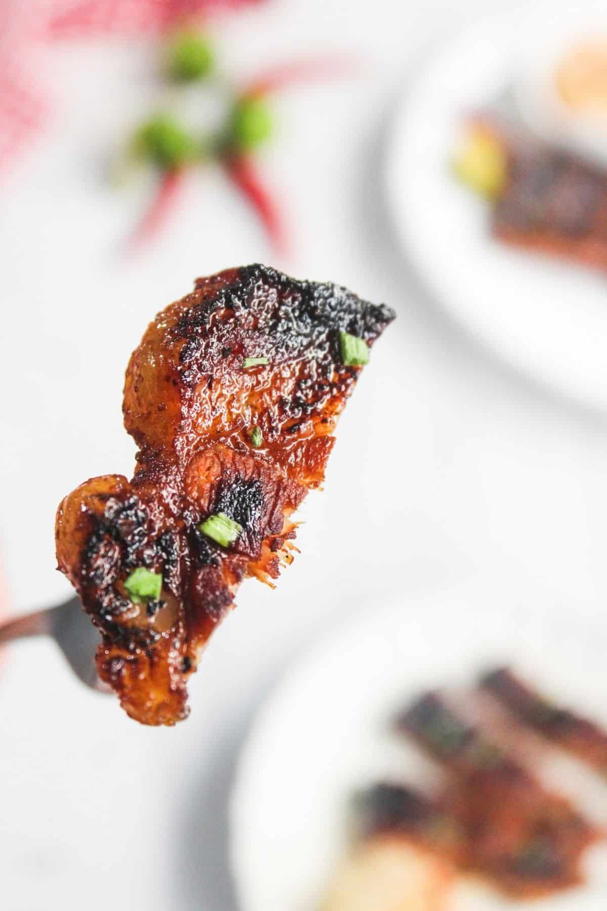 A close-up of a fork holding a piece of grilled, seasoned meat garnished with chopped green herbs, with blurred plates in the background.