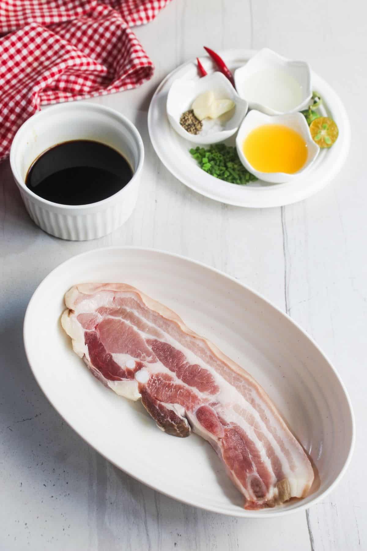 Raw pork belly slices on a white oval plate, with bowls of soy sauce, oil, and seasonings on a separate dish in the background.