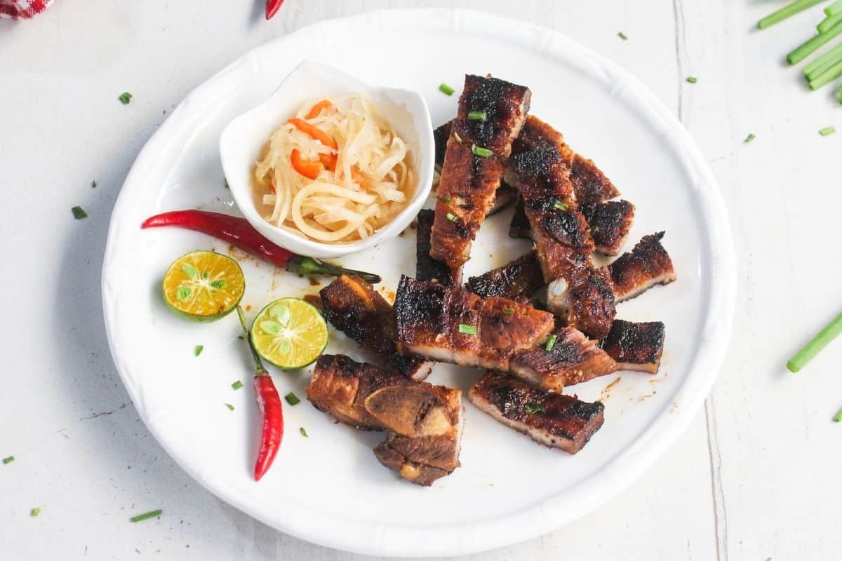 Grilled meat slices arranged on a white plate with calamansi, red chili peppers, and a small bowl of pickled vegetables.