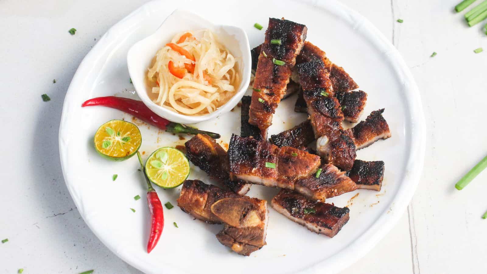 Grilled, sliced meat served on a white plate with halved calamansi, red chili peppers, and a small bowl of pickled vegetables.