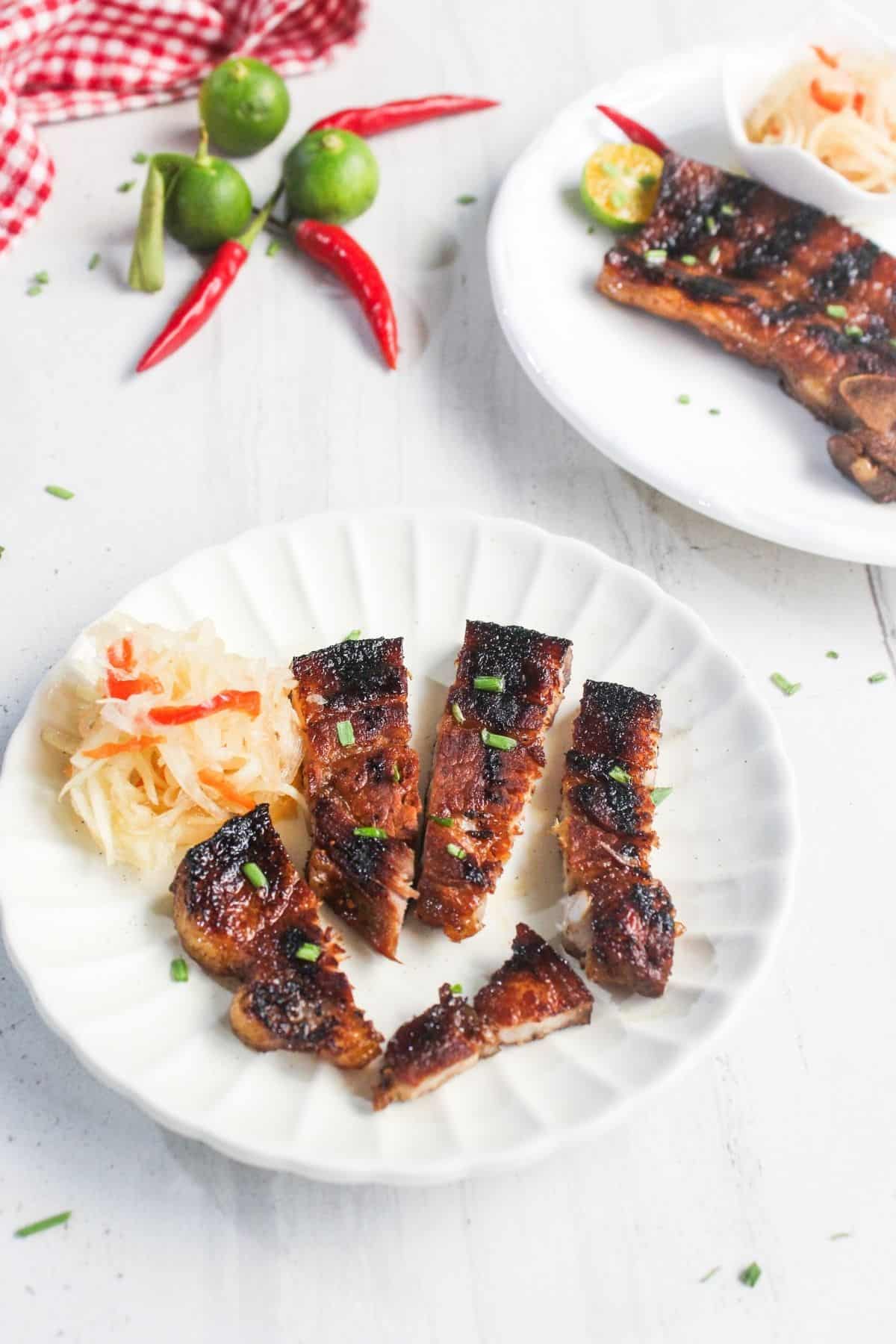 Sliced grilled meat with a side of pickled vegetables on a white plate, garnished with chopped chives; red chilies and calamansi in the background.
