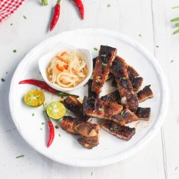 Grilled pork slices on a white plate with a side of pickled vegetables, sliced calamansi, and red chilies, garnished with chopped herbs.