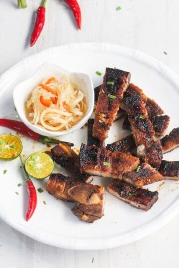 Grilled pork slices on a white plate with a side of pickled vegetables, sliced calamansi, and red chilies, garnished with chopped herbs.