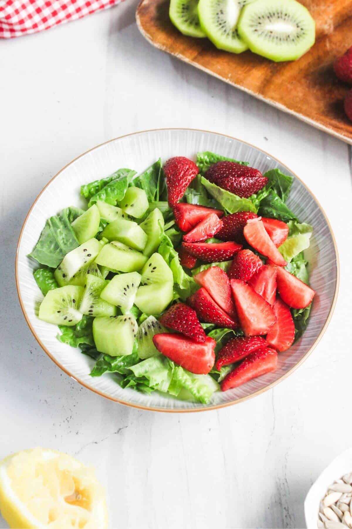 A bowl of salad with chopped kiwi, sliced strawberries, and green leafy lettuce on a white surface.