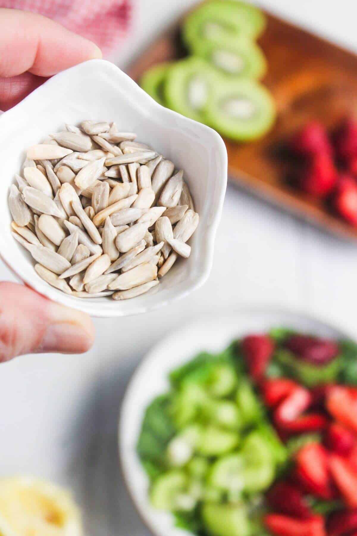 A hand holds a small white bowl filled with sunflower seeds, with sliced kiwi, strawberries, and greens blurred in the background.