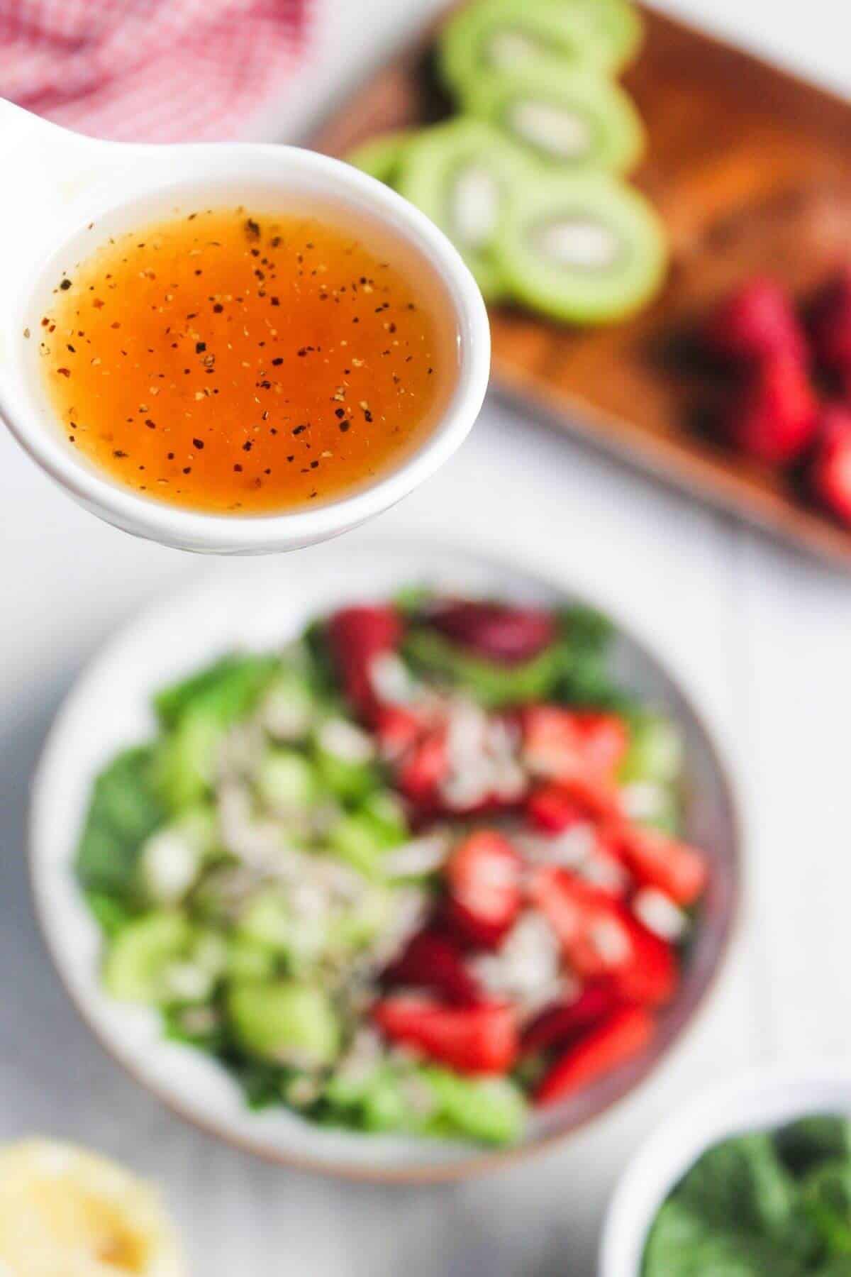 A close-up of a spoonful of vinaigrette dressing held above a salad bowl, with sliced kiwi and strawberries on a board in the background.