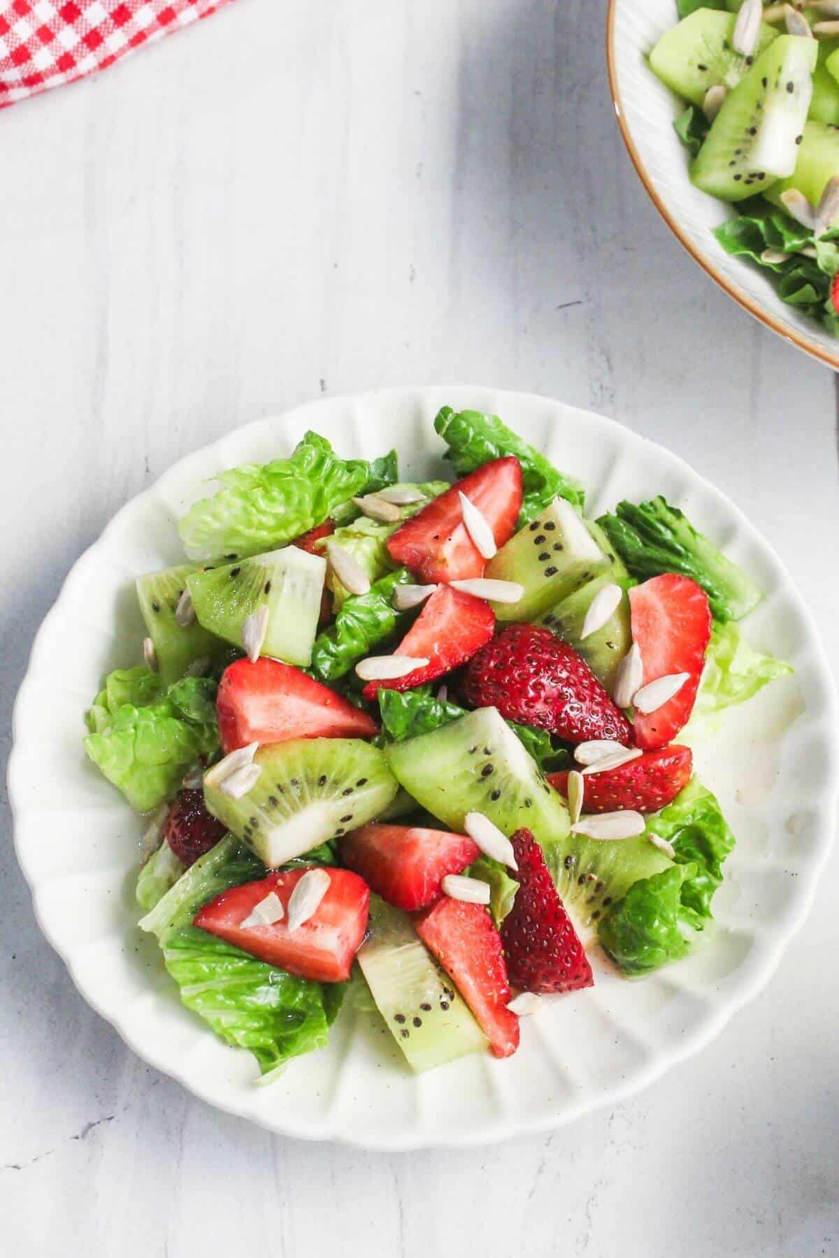 A white plate with a salad made of lettuce, sliced strawberries, kiwi, and sunflower seeds, set on a light surface.
