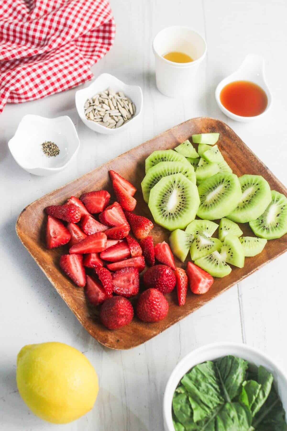 A wooden plate with sliced kiwi and strawberries next to a lemon, spinach, seeds, black pepper, and small cups of liquids on a white surface.