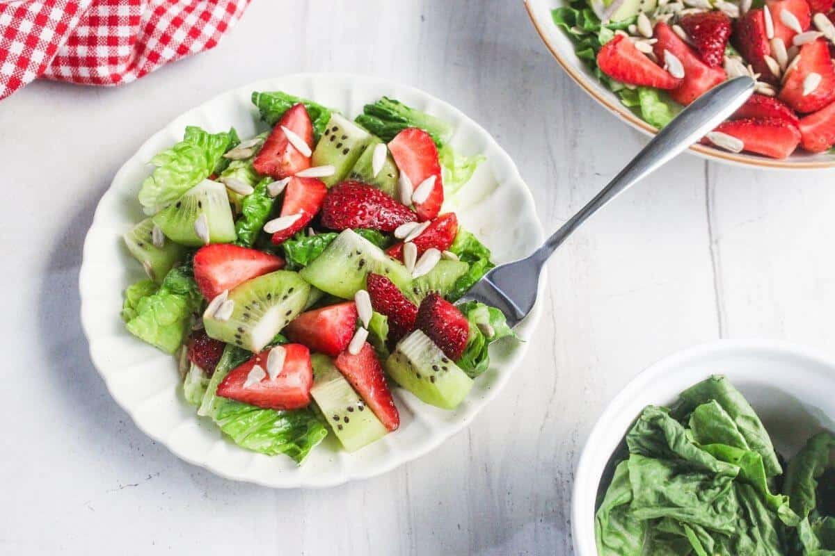 A white plate with a salad of lettuce, sliced kiwi, strawberries, and slivered almonds, with a fork on the side and extra greens in nearby bowls.