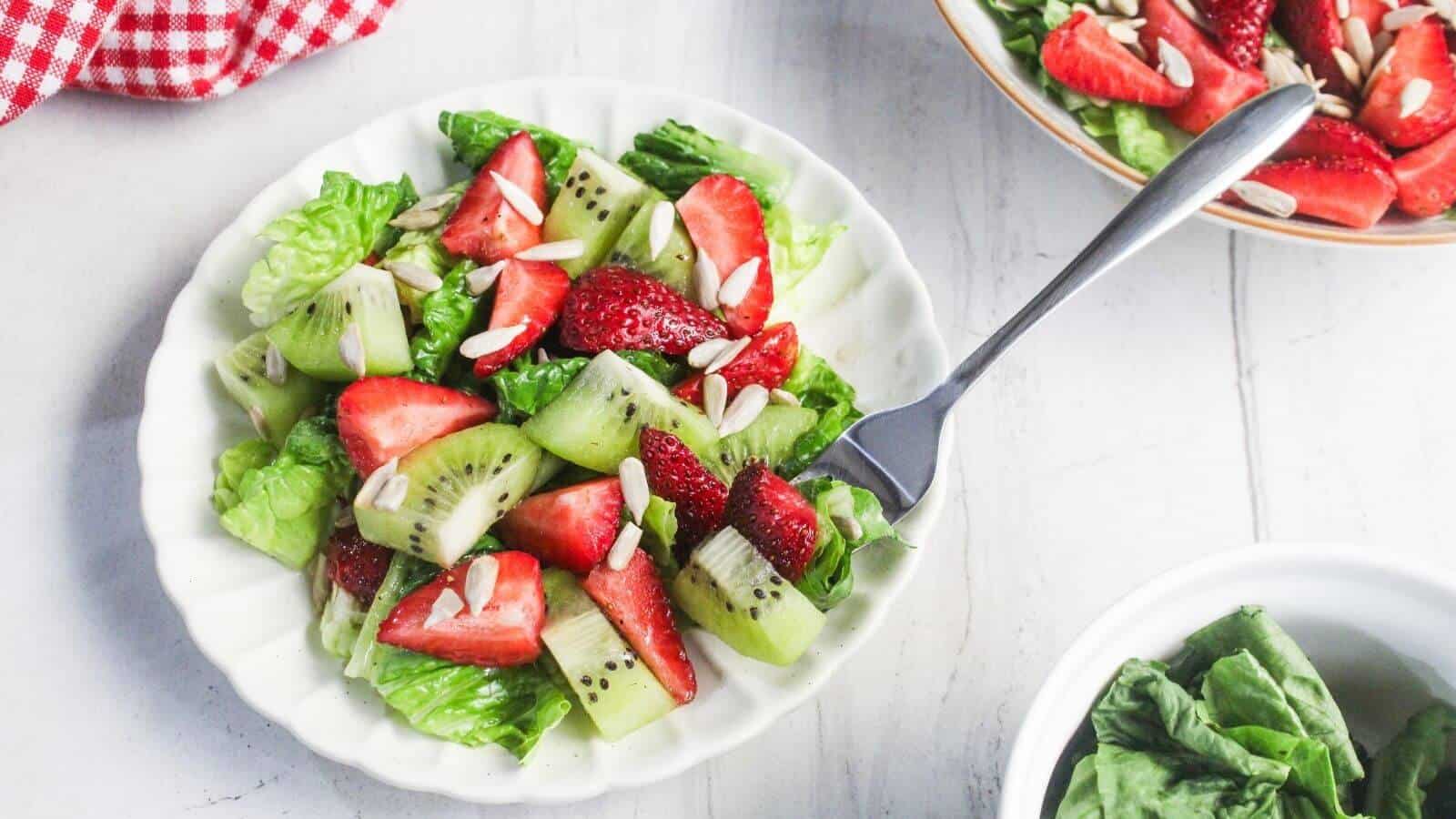 A white plate with a salad of lettuce, sliced strawberries, kiwi, and slivered almonds, with a fork resting on the plate.