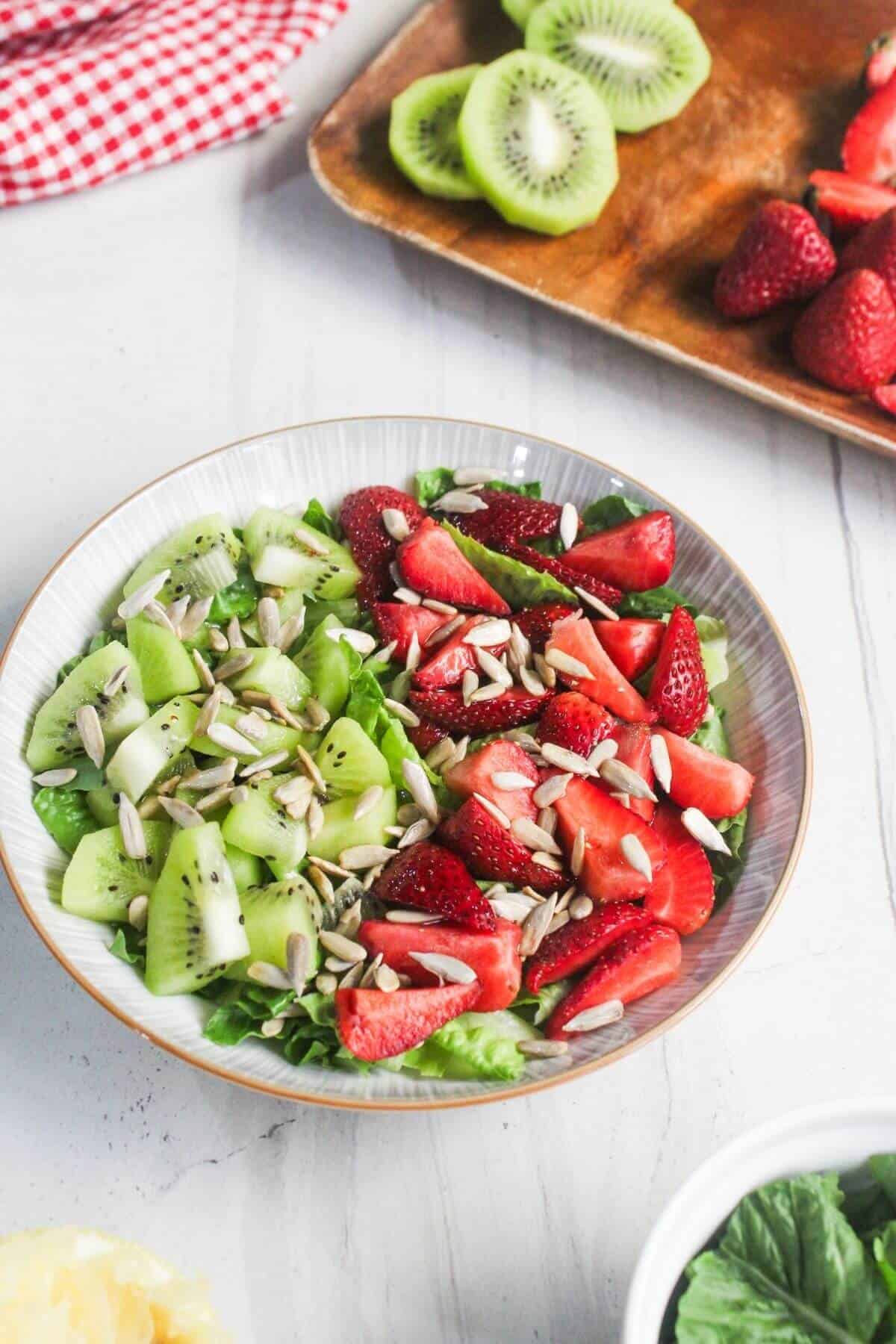 A bowl of salad topped with sliced kiwi, strawberries, and sunflower seeds on a white surface, with extra fruit on a tray in the background.