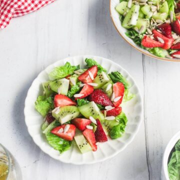 A plate of salad with lettuce, sliced strawberries, kiwi, cucumber, and slivered almonds on a white table beside a bowl of the same salad.
