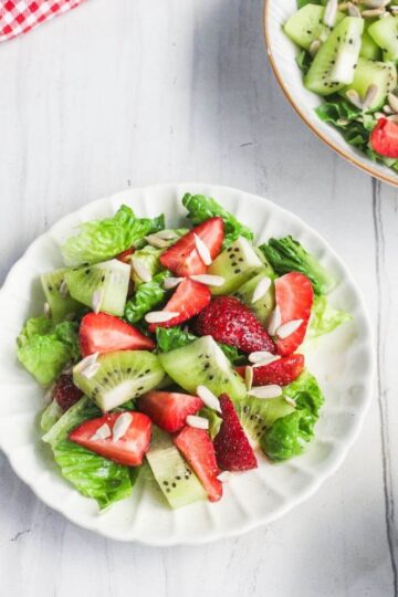 A plate of salad with lettuce, sliced strawberries, kiwi, cucumber, and slivered almonds on a white table beside a bowl of the same salad.