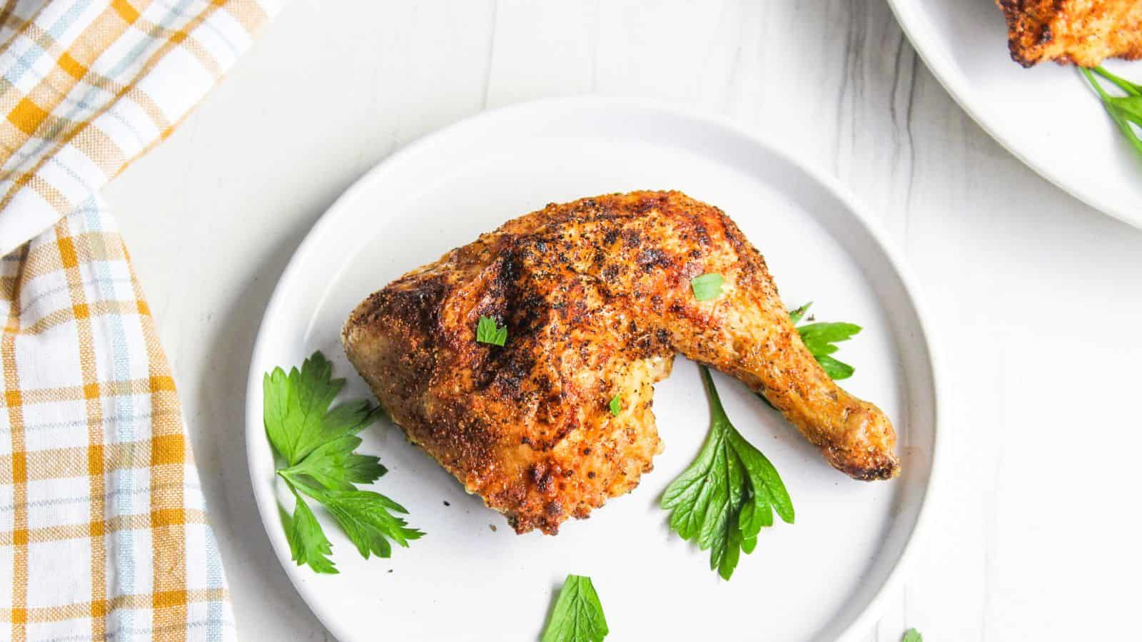 A cooked, seasoned chicken leg quarter is served on a white plate with parsley garnish. A checkered napkin and part of another plate are visible nearby.