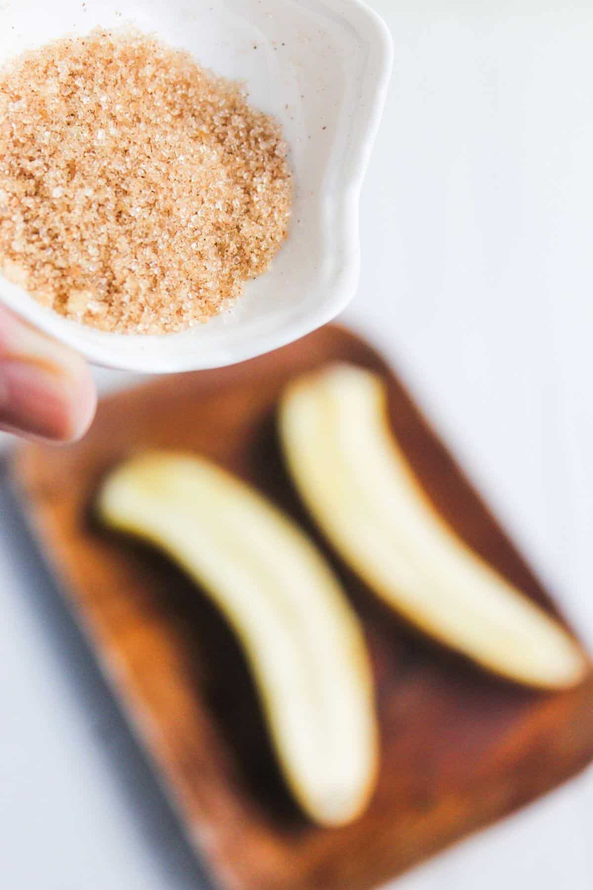 A small bowl of cinnamon sugar held above a wooden board with two peeled bananas.