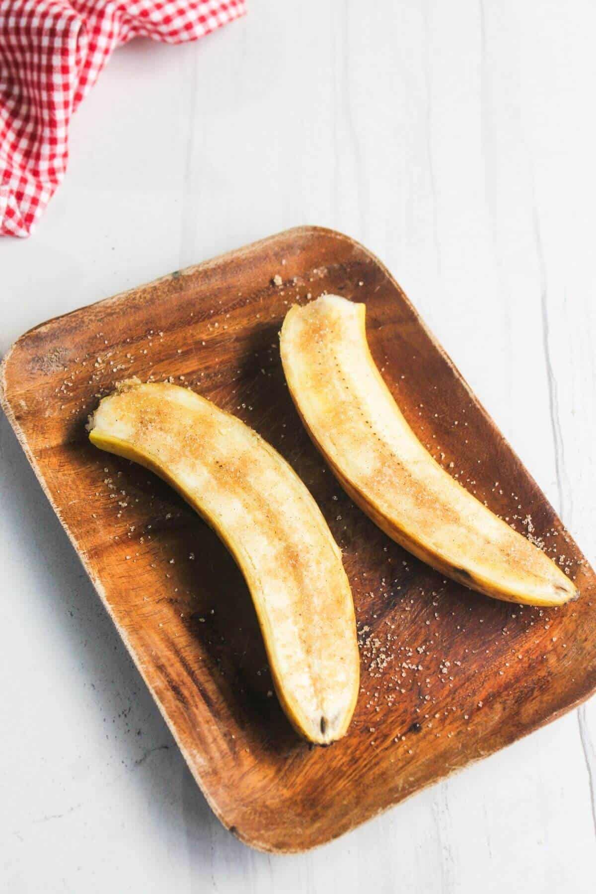 Two banana halves sprinkled with cinnamon and sugar on a wooden plate, set on a light-colored surface with a red and white checkered cloth in the corner.