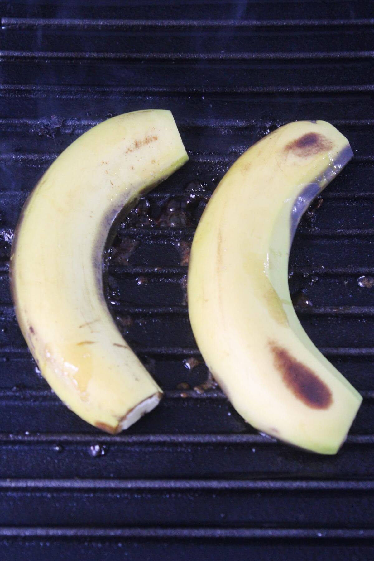 Two unpeeled bananas with slight brown spots cooking on a ridged grill pan.
