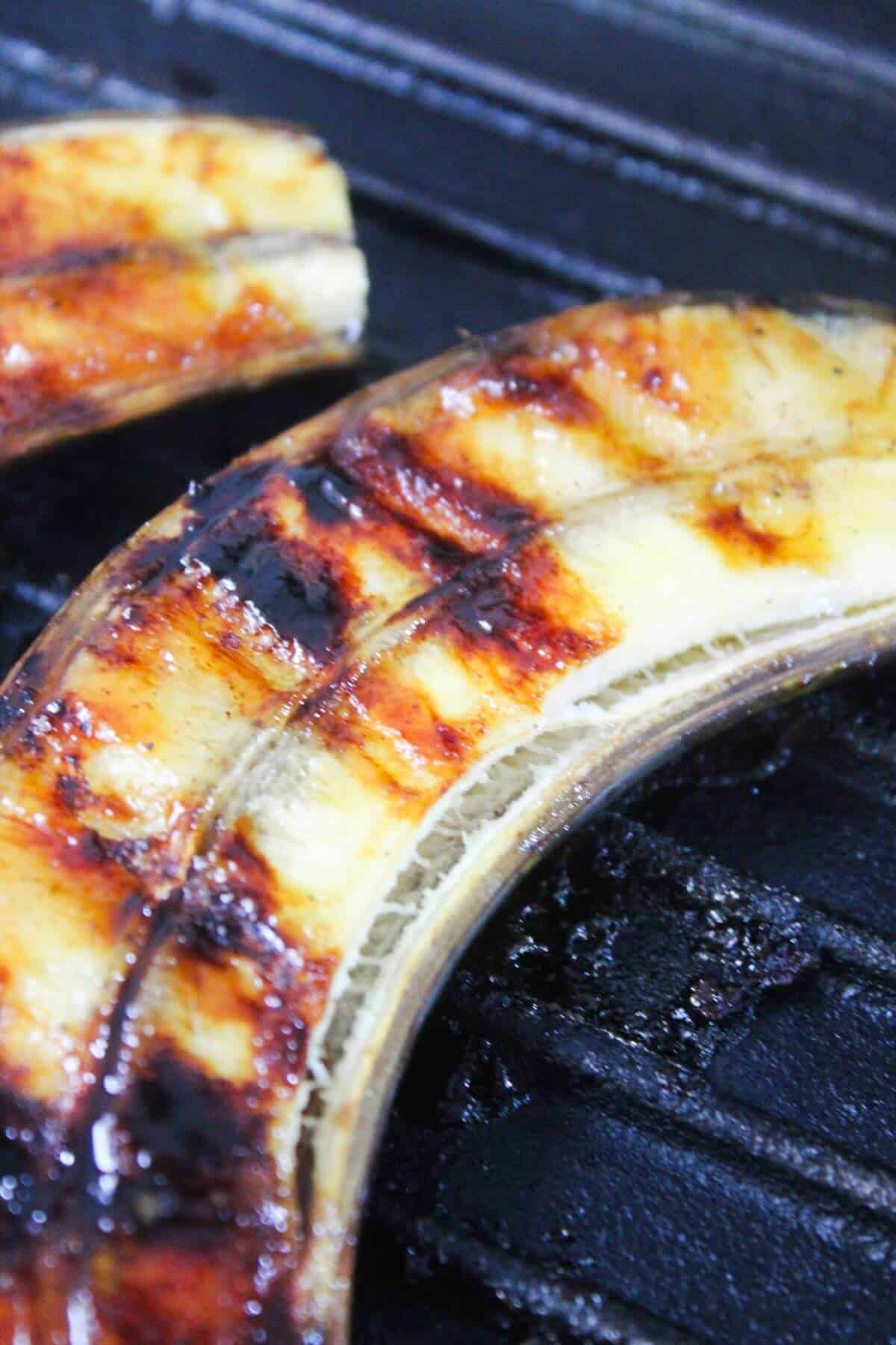 Close-up of banana halves with peel on, grilling on a dark grill surface, showing visible grill marks and caramelization.