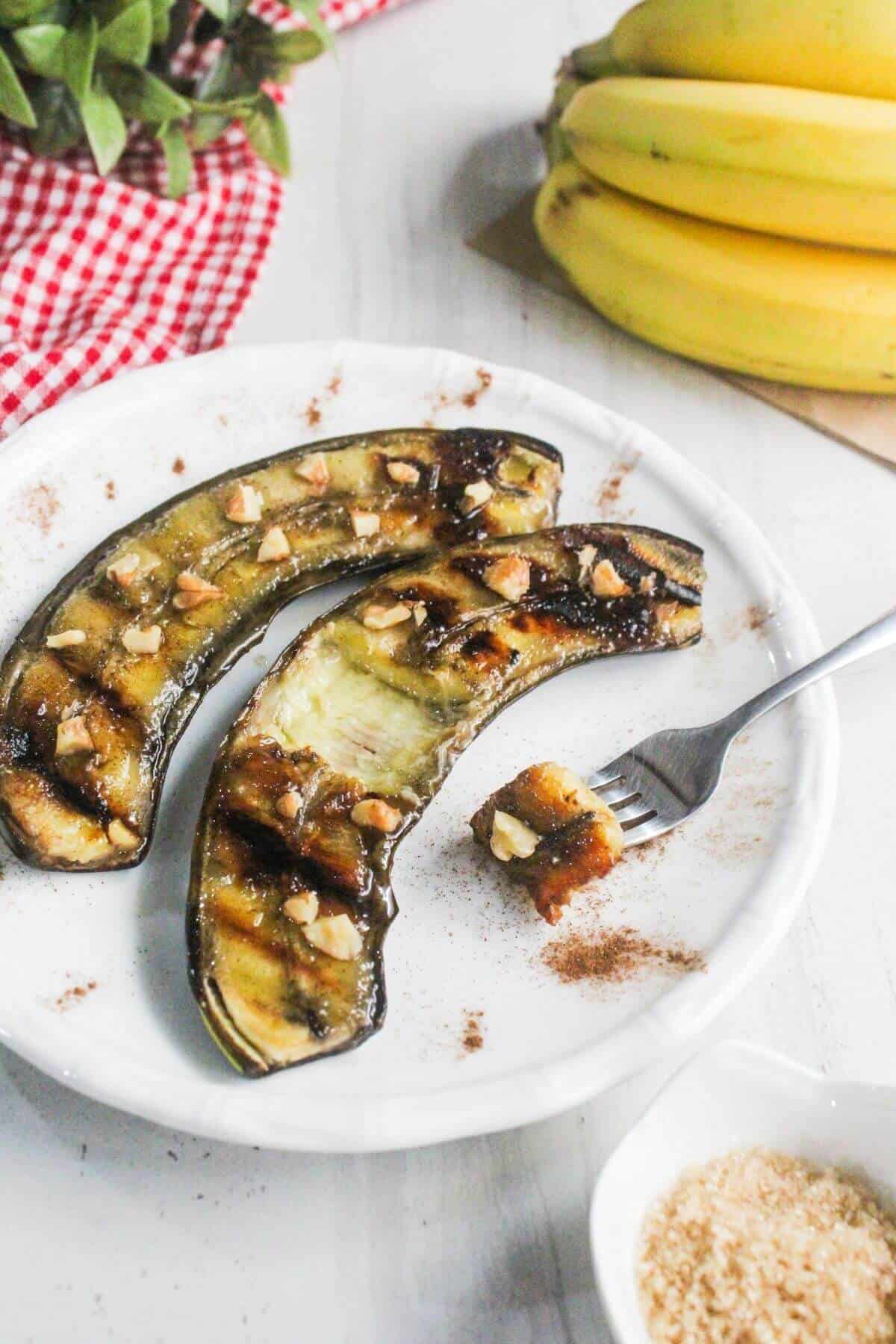 Two baked bananas topped with chopped nuts on a white plate, with a fork holding a bite. A bunch of bananas and a red checkered cloth are in the background.