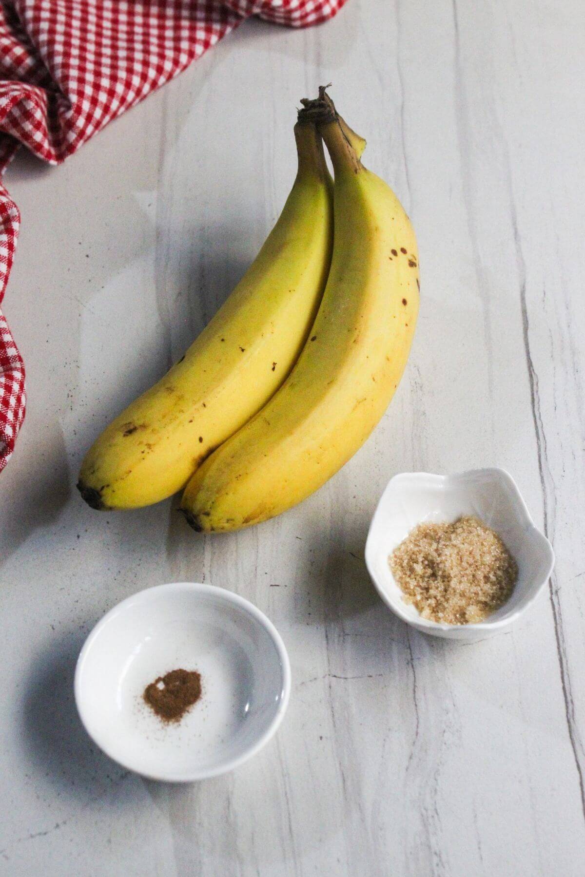 Two ripe bananas, a small bowl of brown sugar, a small bowl of ground cinnamon, and a red checkered cloth on a white marble surface.