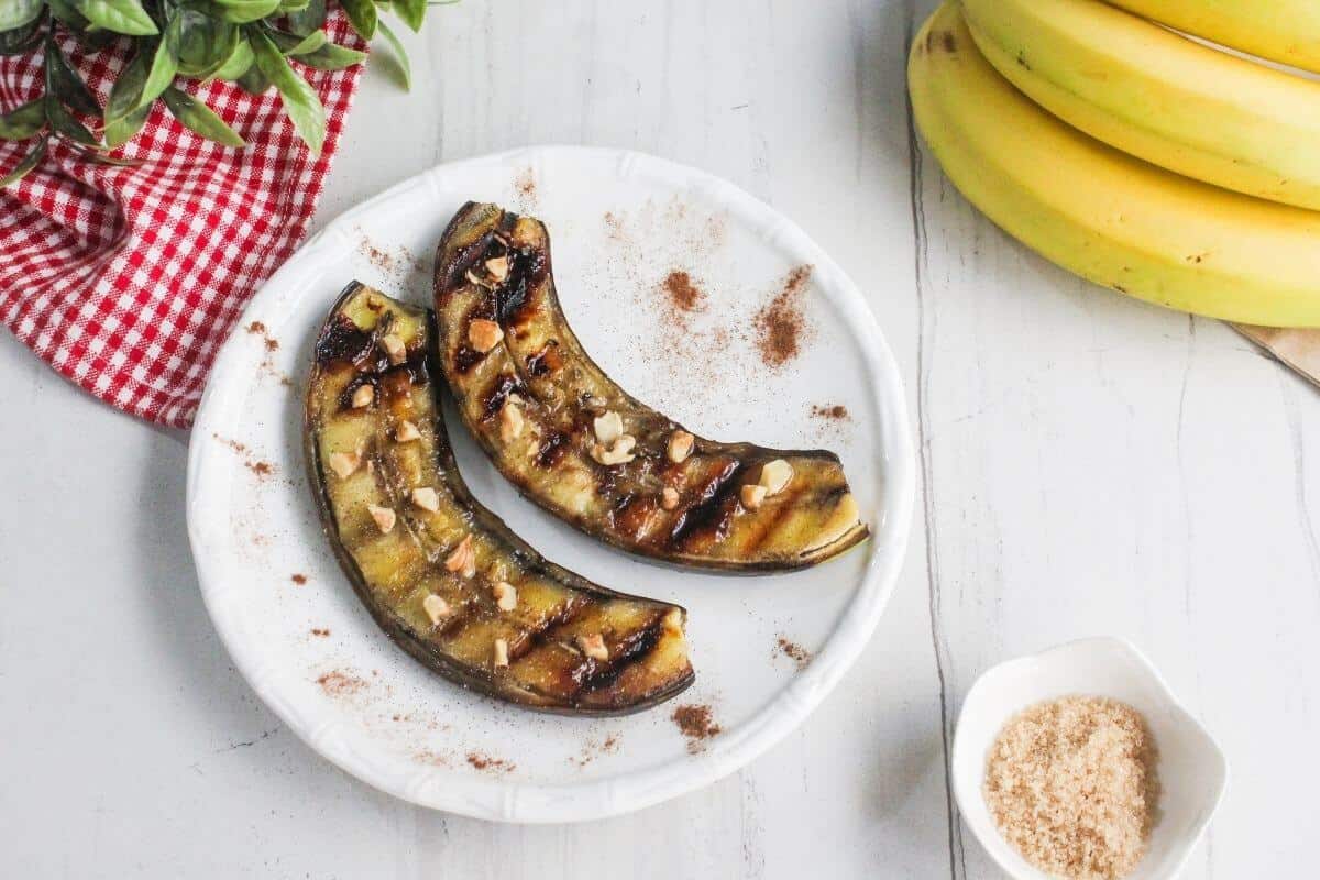 Two grilled banana halves on a white plate, topped with chopped nuts and sprinkled with cinnamon, next to a bunch of bananas and a small bowl of brown sugar.