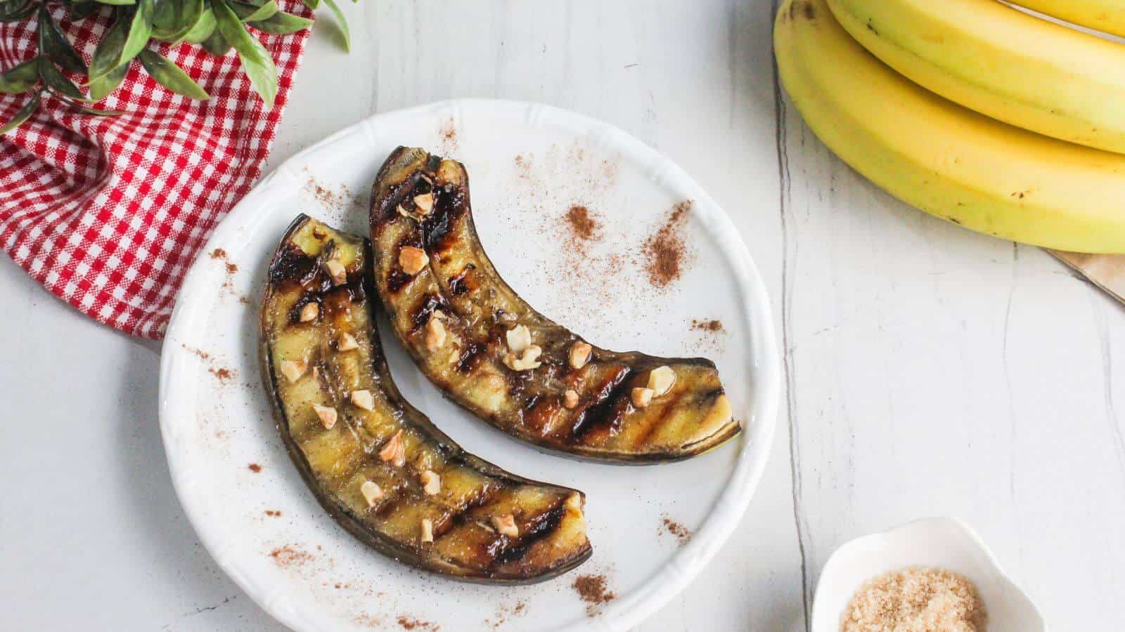 Two grilled banana halves topped with chopped nuts and cinnamon on a white plate, with bananas, a red-checked cloth, and a bowl of sugar nearby.
