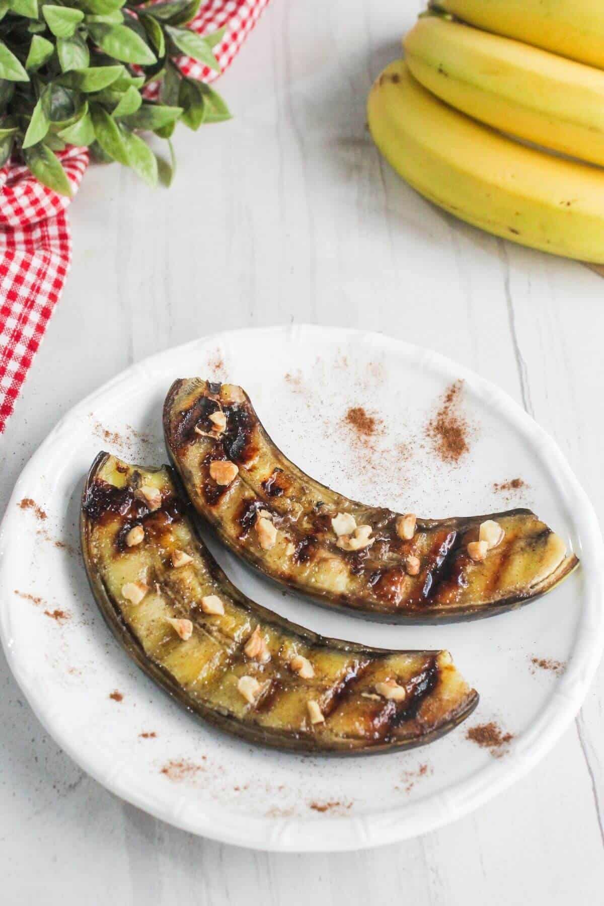 Two grilled bananas topped with chopped nuts and drizzled sauce on a white plate, with a red checkered cloth and bananas in the background.