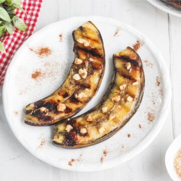 Two grilled banana halves topped with chopped nuts are arranged on a white plate, with a sprinkle of cinnamon on the surface.