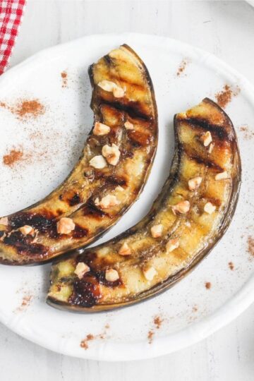 Two grilled banana halves topped with chopped nuts are arranged on a white plate, with a sprinkle of cinnamon on the surface.