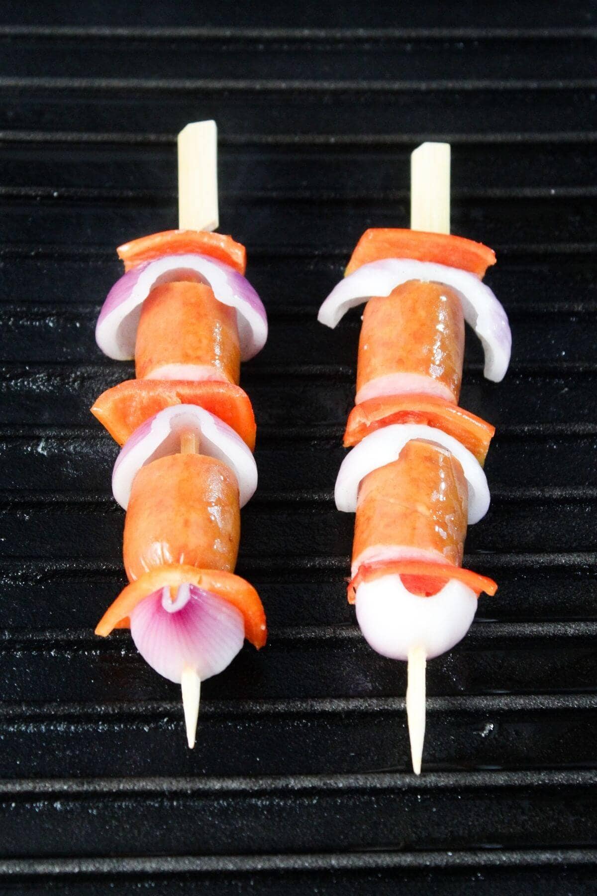 Two skewers with sliced sausage, red onion, and red bell pepper are placed on a black grill surface.