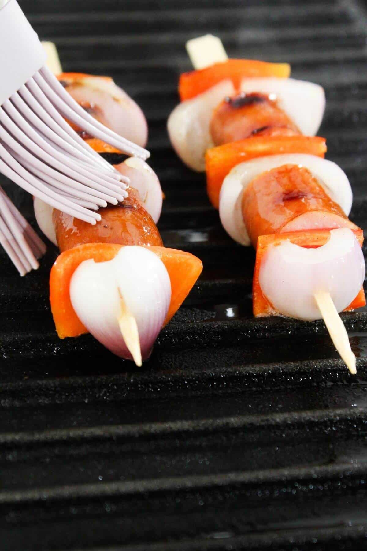 Vegetable and sausage skewers are brushed with sauce while cooking on a grill.