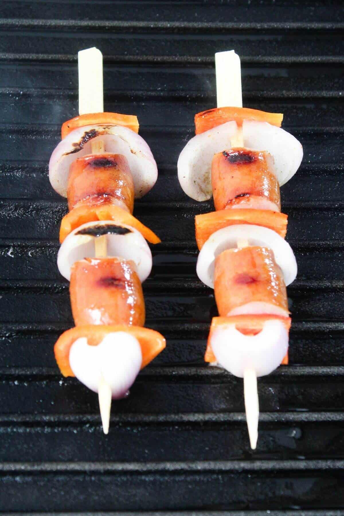 Two skewers with sausage, bell pepper, and onion slices are cooking on a grill.