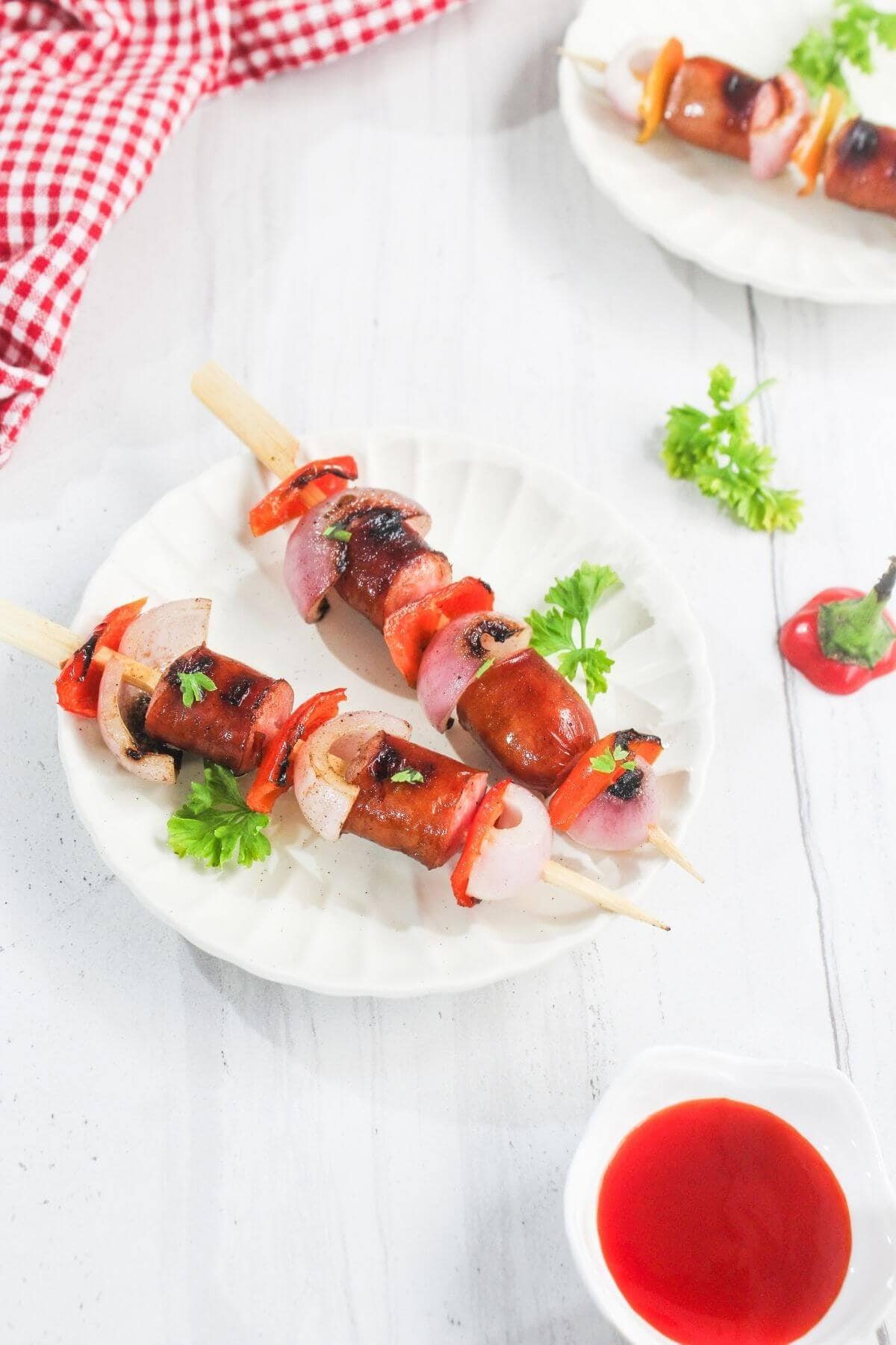 Grilled sausage and vegetable skewers on a white plate, garnished with parsley, with a small bowl of red sauce nearby.