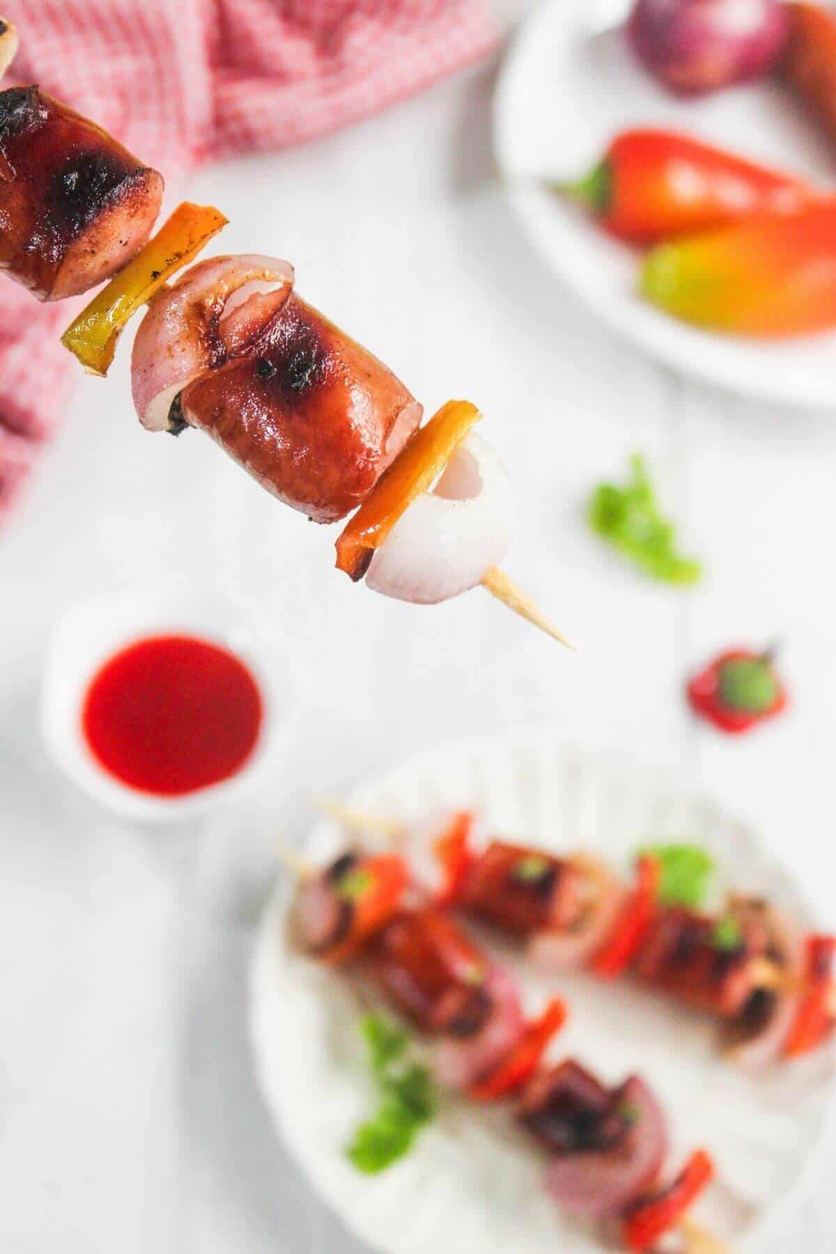 A close-up of a grilled sausage and vegetable skewer, featuring onions and peppers, with more skewers, sauce, and peppers blurred in the background.