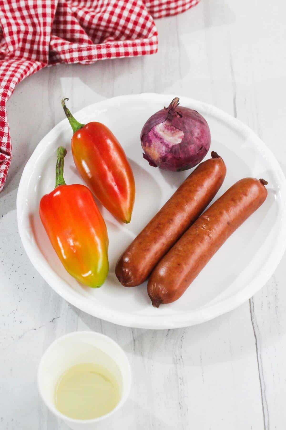 Two sausages, two bell peppers, and a red onion on a white plate, with a small cup of oil and a red checkered cloth nearby on a white surface.