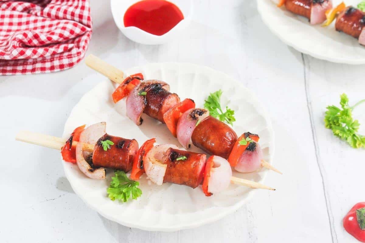 Three grilled sausage and vegetable skewers are served on a white plate with parsley garnish, next to a dish of red sauce and a red checkered cloth.