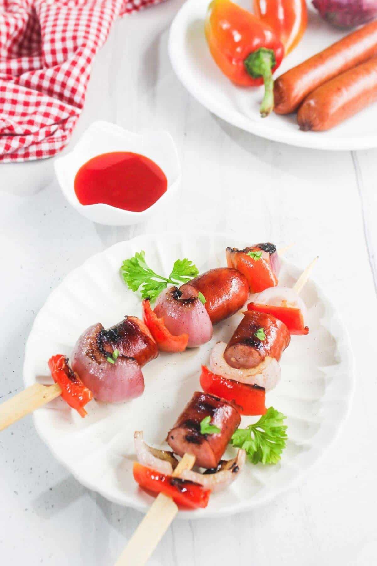 Two grilled skewers with sausage, red bell pepper, and onion on a white plate, garnished with parsley. A small bowl of red sauce and vegetables are in the background.