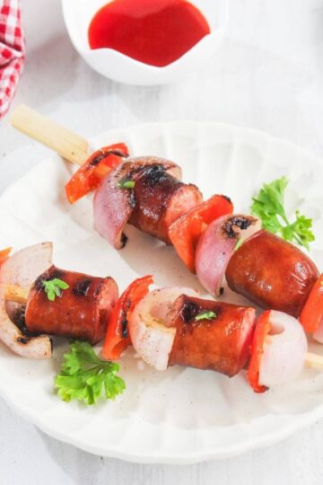 Two skewers with grilled sausage, red bell pepper, and onion are served on a white plate with parsley garnish, next to a bowl of red sauce and a red-checked cloth.