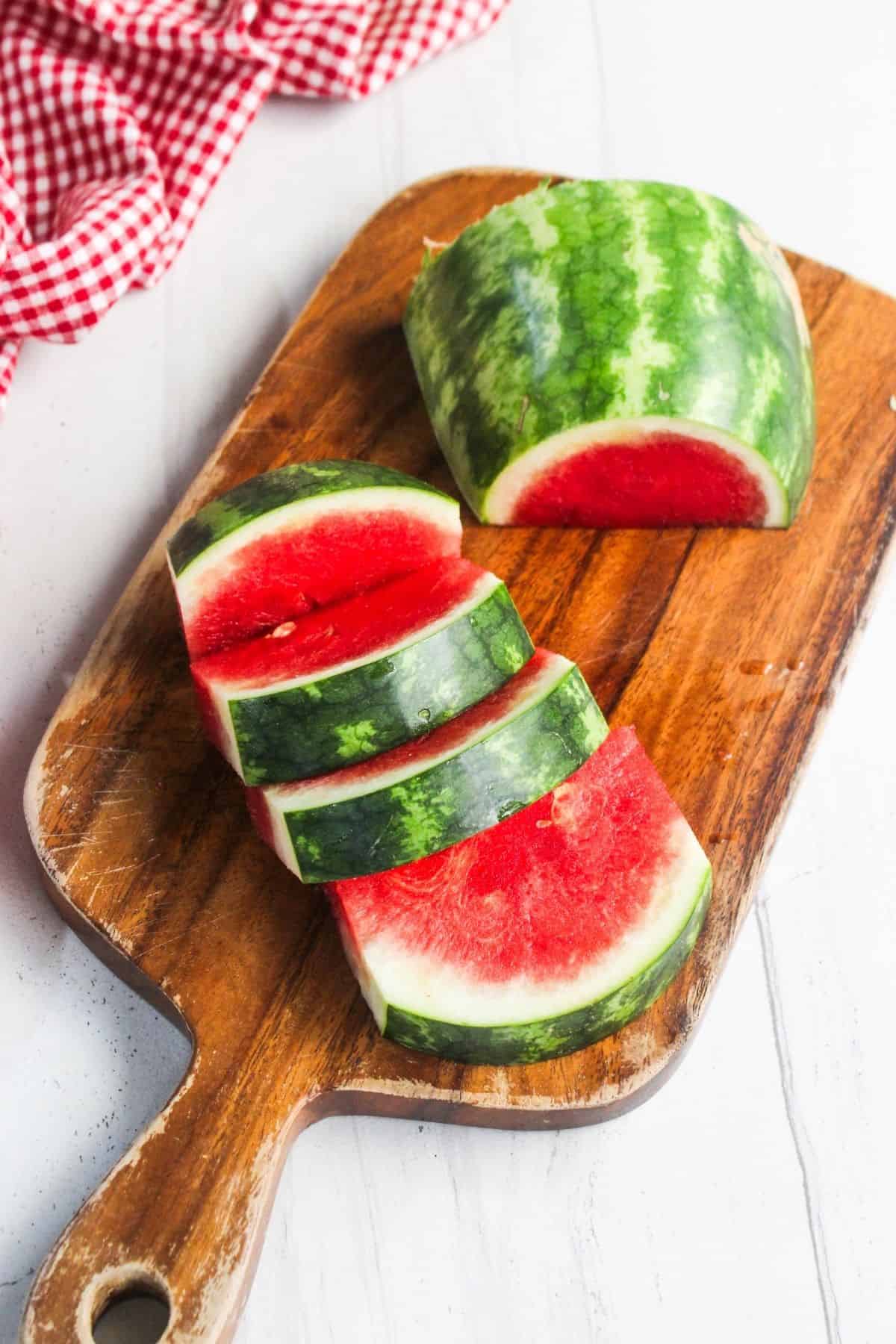 A partially sliced watermelon with three cut pieces on a wooden cutting board, next to a red and white checkered cloth.