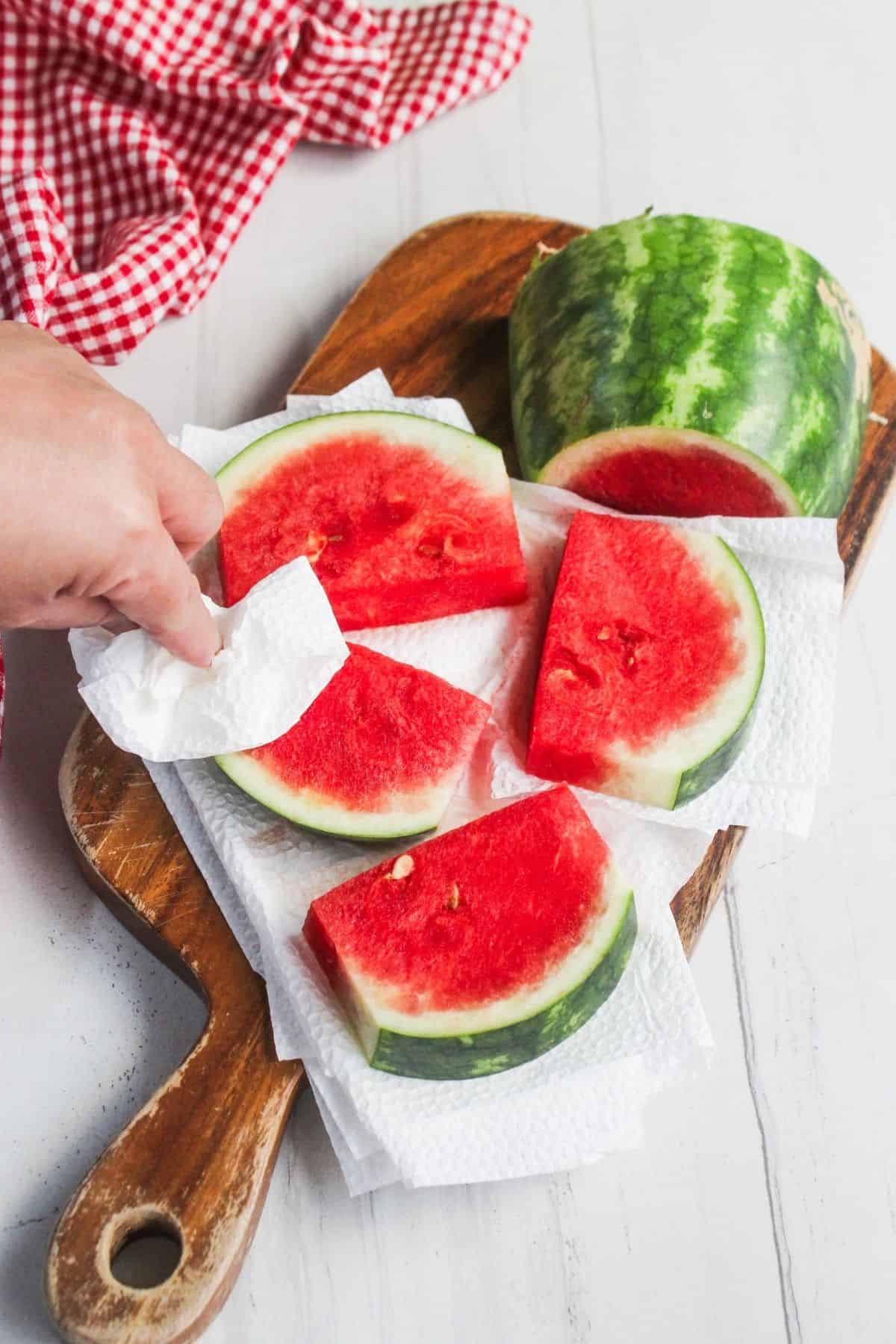 A hand uses a paper towel to pat dry watermelon slices arranged on a wooden cutting board with a red gingham cloth nearby.