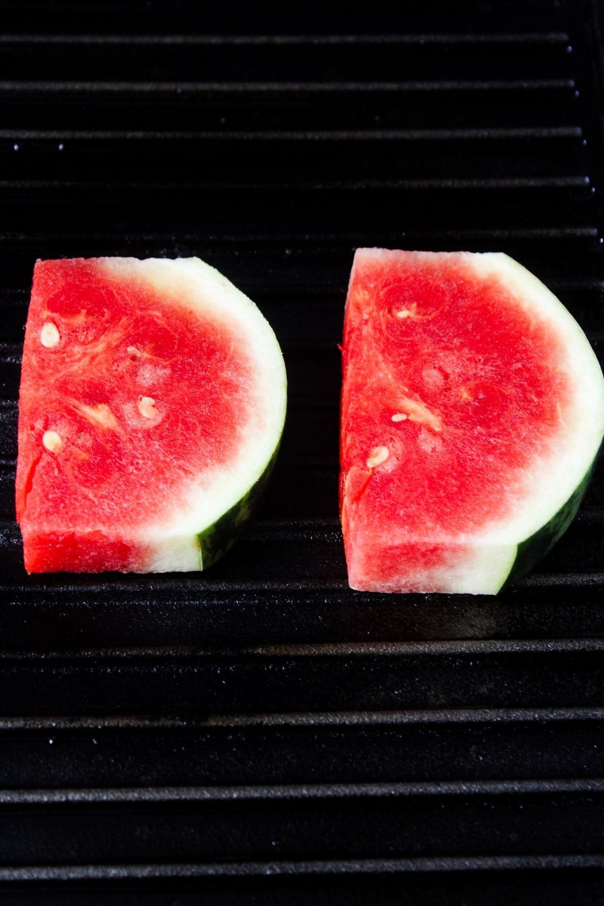 Two slices of watermelon with rind are placed side by side on a black grill.