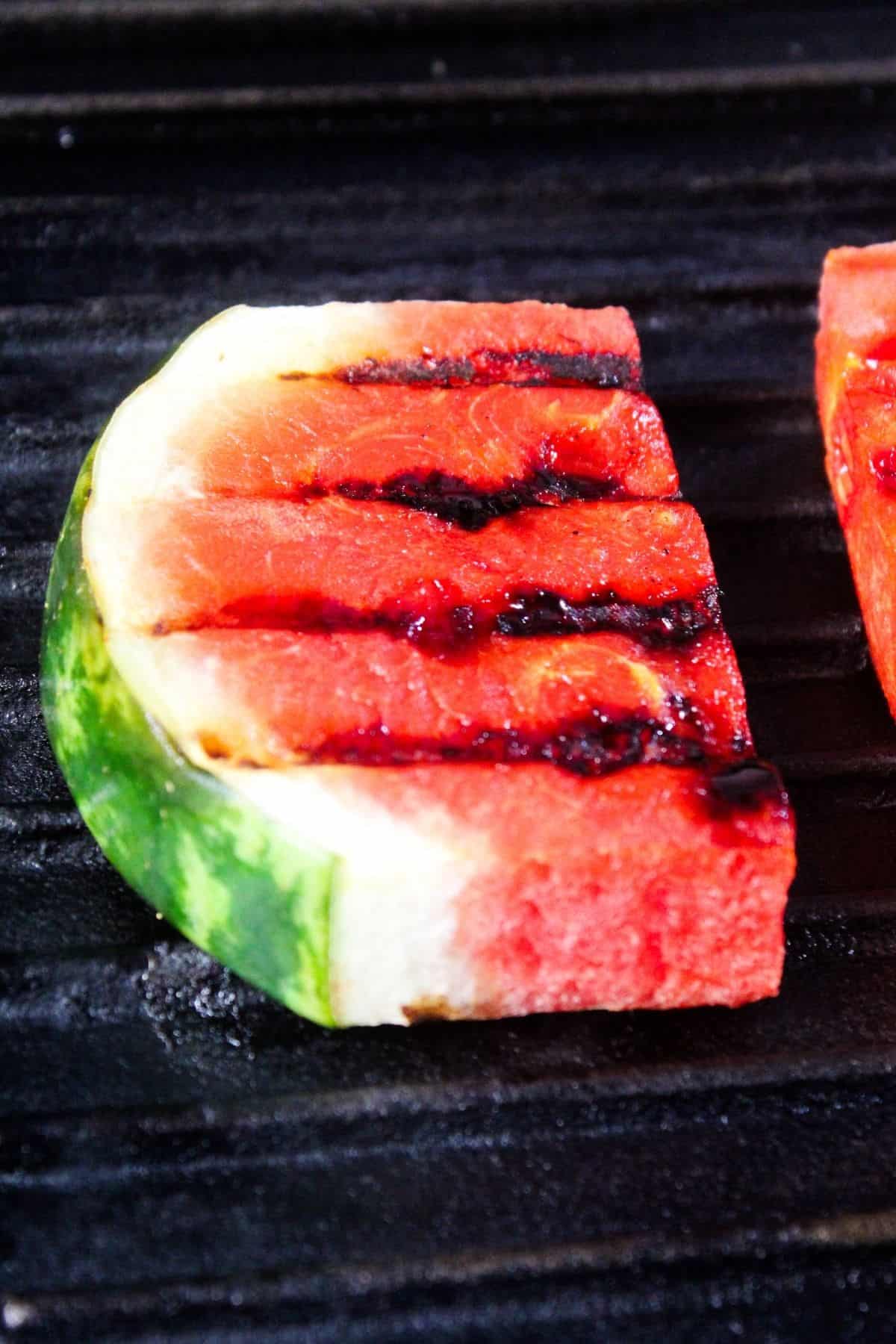 A slice of watermelon with grill marks on its surface sits on a black grill grate.