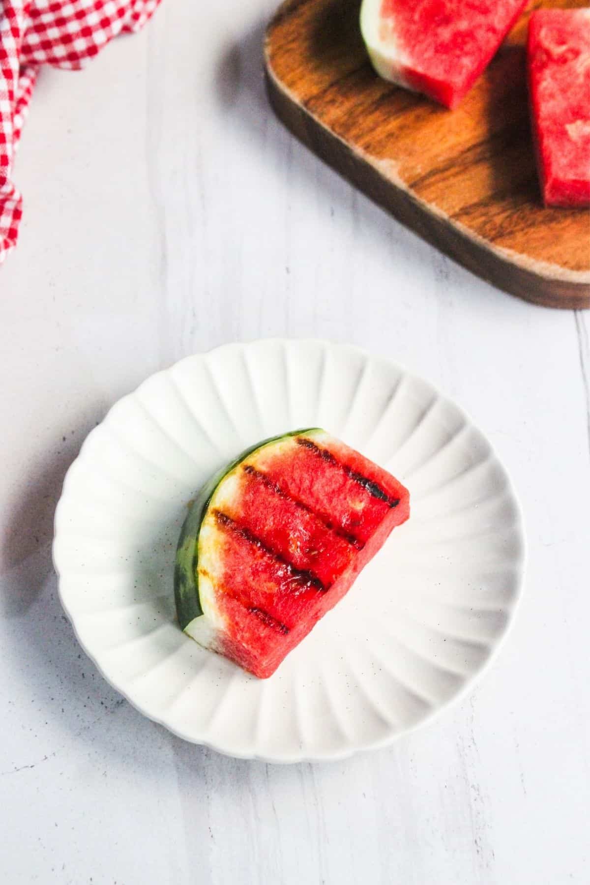 A grilled watermelon wedge sits on a white plate, with a wooden board holding more watermelon slices in the background.