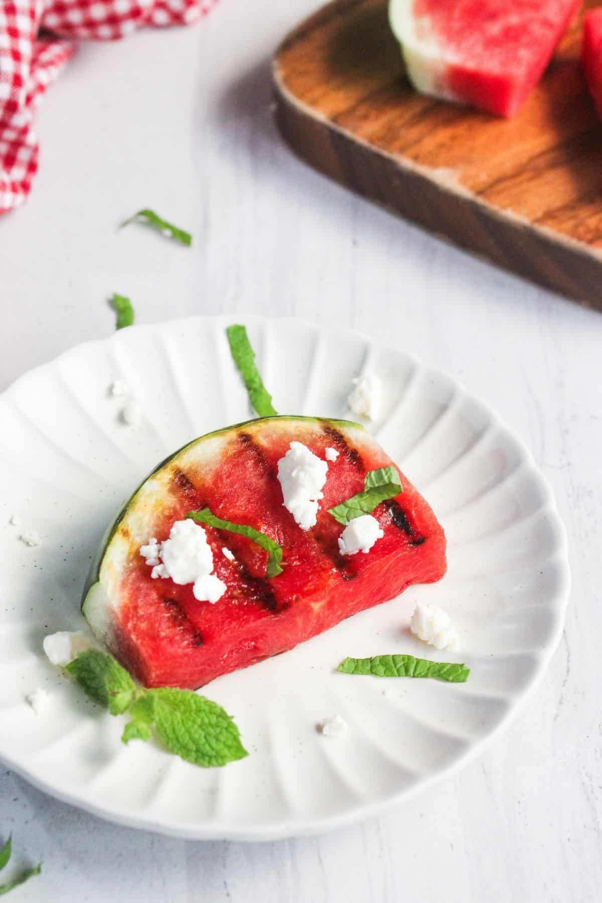 A grilled watermelon slice topped with crumbled cheese and mint leaves sits on a white plate, with more watermelon on a wooden board in the background.