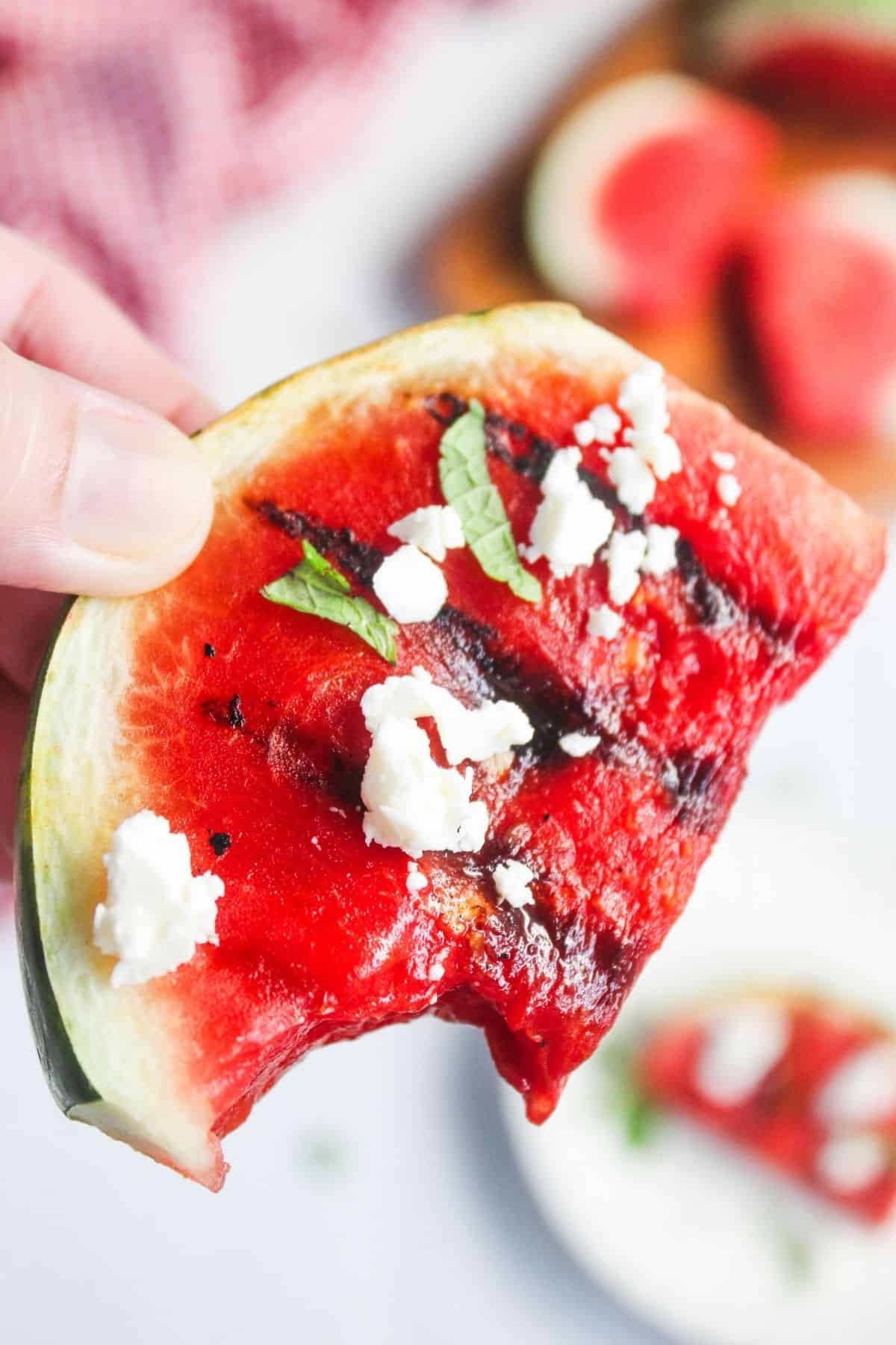 Hand holding a grilled watermelon slice topped with crumbled feta cheese and fresh mint, with a bite taken out.
